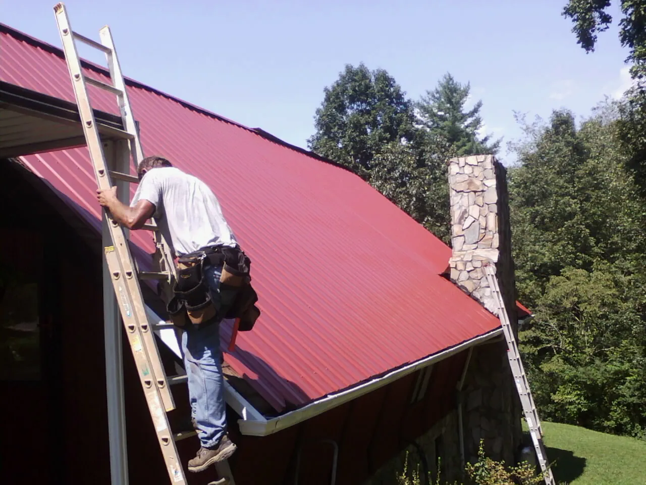 Roofer on a ladder beside a red metal roof and stone chimney — Soffit & Fascia Repair by Roofing Brothers in North Norwich